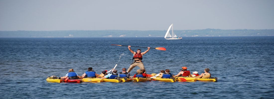 Brody-standing-on-kayaks-with-sailboat-behind-1920×1080 Kayaks with a sailboat behind them in Door County, Wisconsin